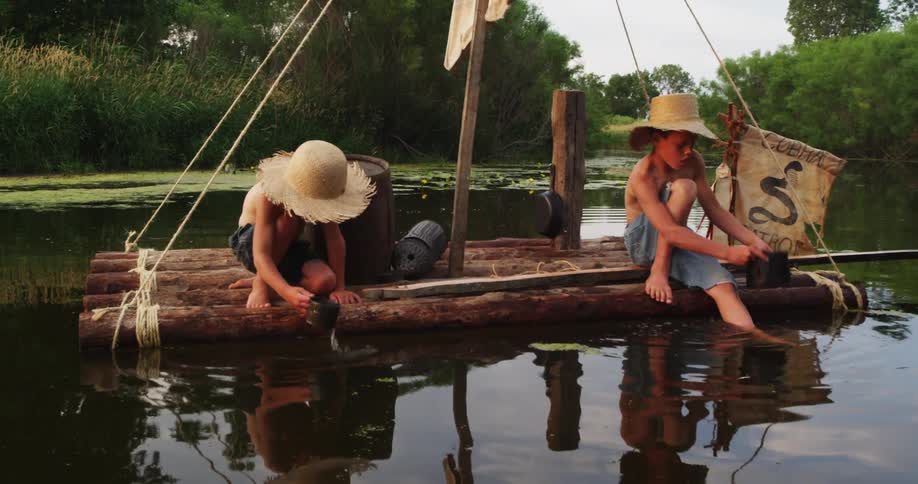 A pair of boys sitting on a wooden raft in a marsh scooping water with tin pots