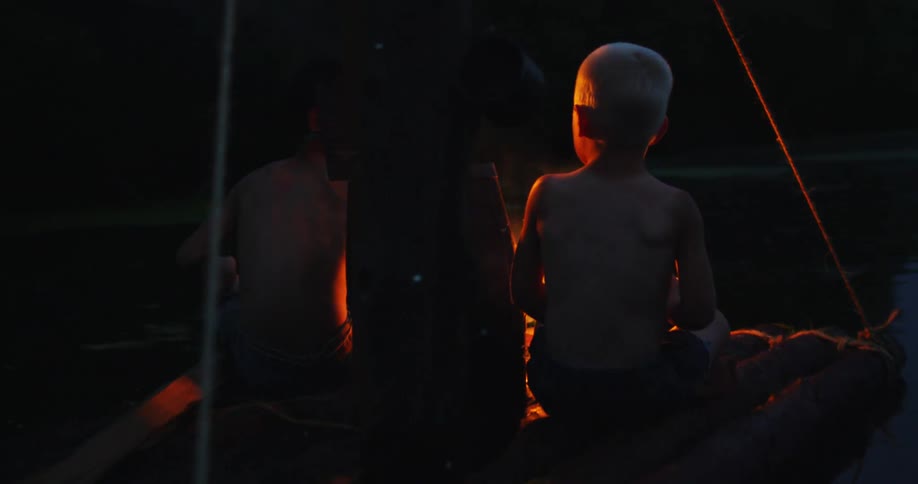 Two young boys eating next to small fire on wooden raft on lake