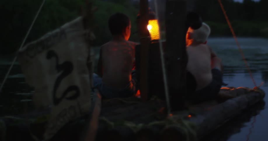 Two boys sit on a raft floating in a lake with a fire going and a flag banner on the back