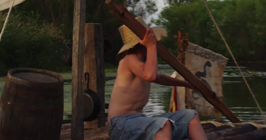 Two young boys ride a raft made of wooden logs down a river