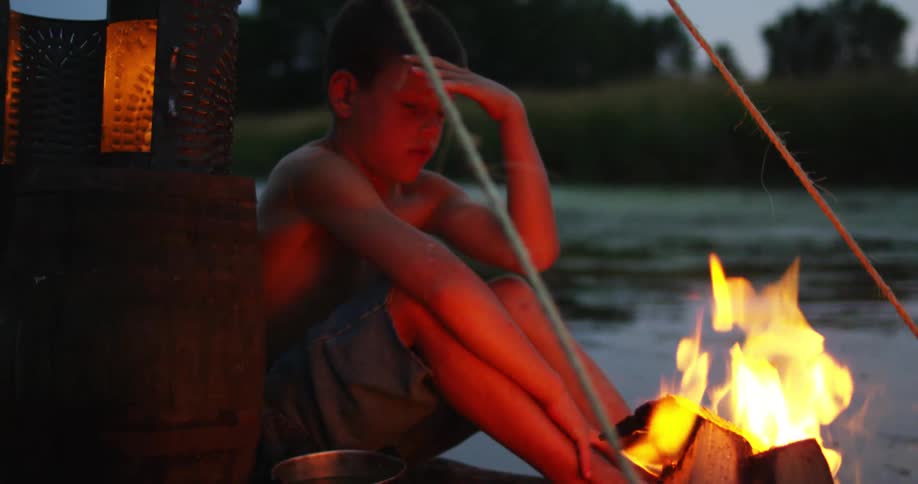 Boy sitting in front of campfire near lake at sunset
