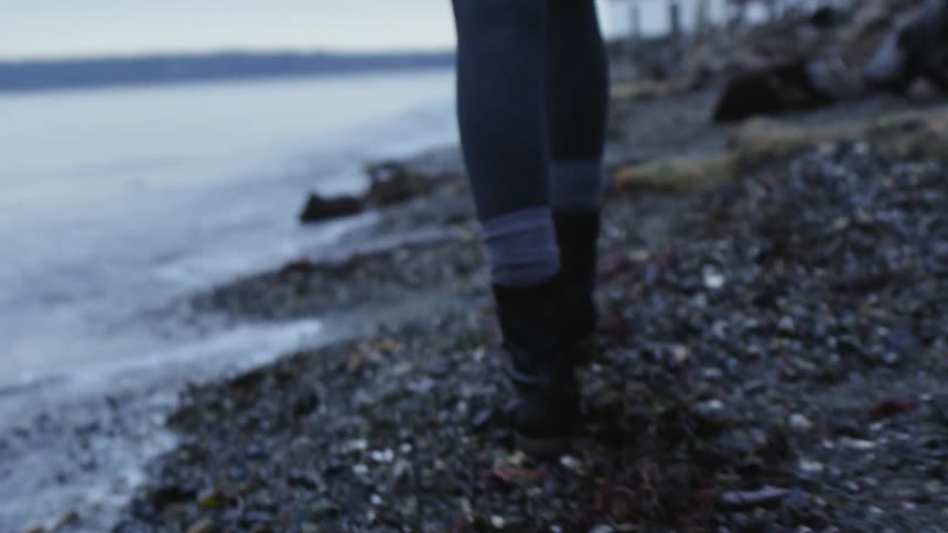 A woman in boots walks along a rocky shoreline