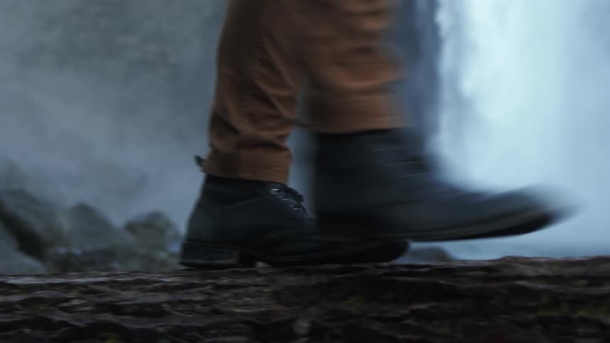 A person wearing heavy boots carefully walks across a log by a waterfall