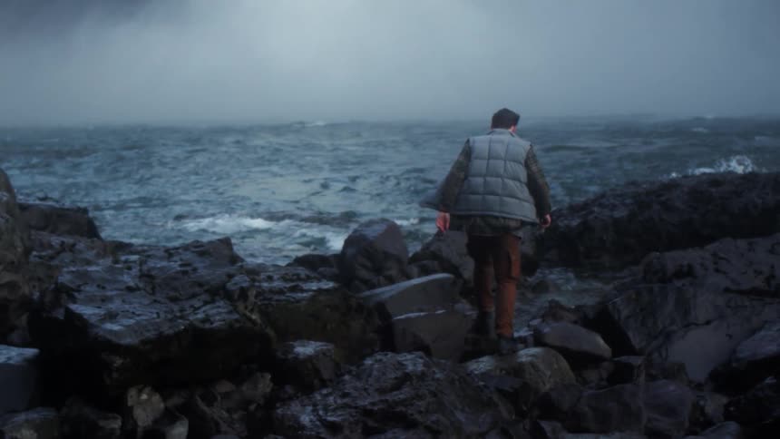 A white man carefully walks on rocks next to a body of water fed by an enormous waterfall
