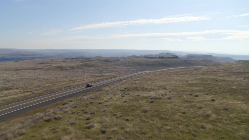 Skimming over a desert landscape as a black car drives along an empty two lane road