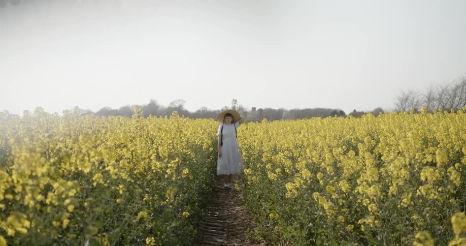 A young woman in a sun hat standing on narrow dirt path through a field of tall yellow flowers