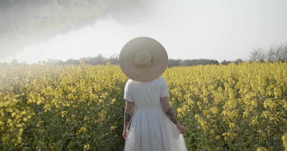 A woman in a white dress and sunhat walks in a field of yellow flowers