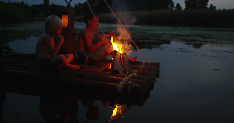 Two boys on a raft at night sit around a fire eating watermelon
