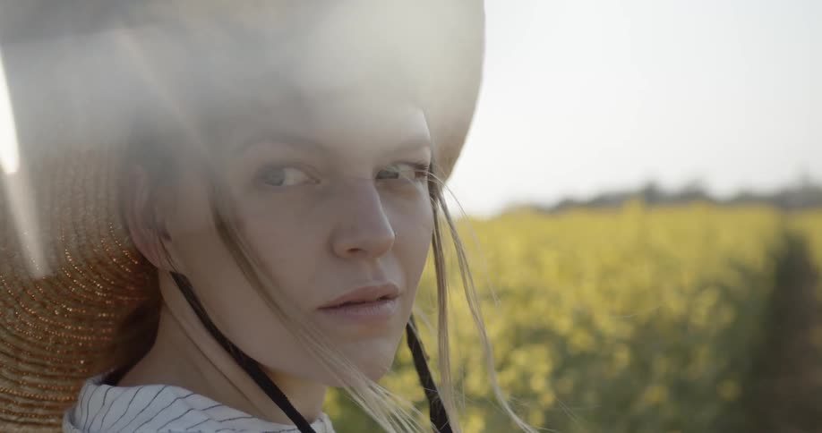 A white woman in a sun hat stands outdoors next to a field of flowers