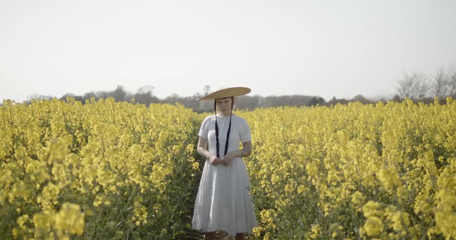 A white woman in a dress and sun hat stands outdoors surrounded by flowers
