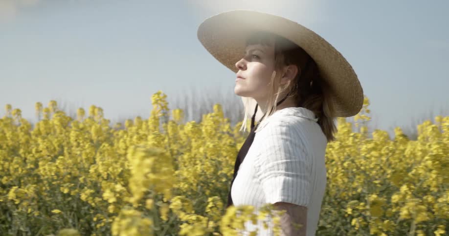 A woman holds her straw hat on her head with one hand as she walks through a field of flowers on a bright sunny day