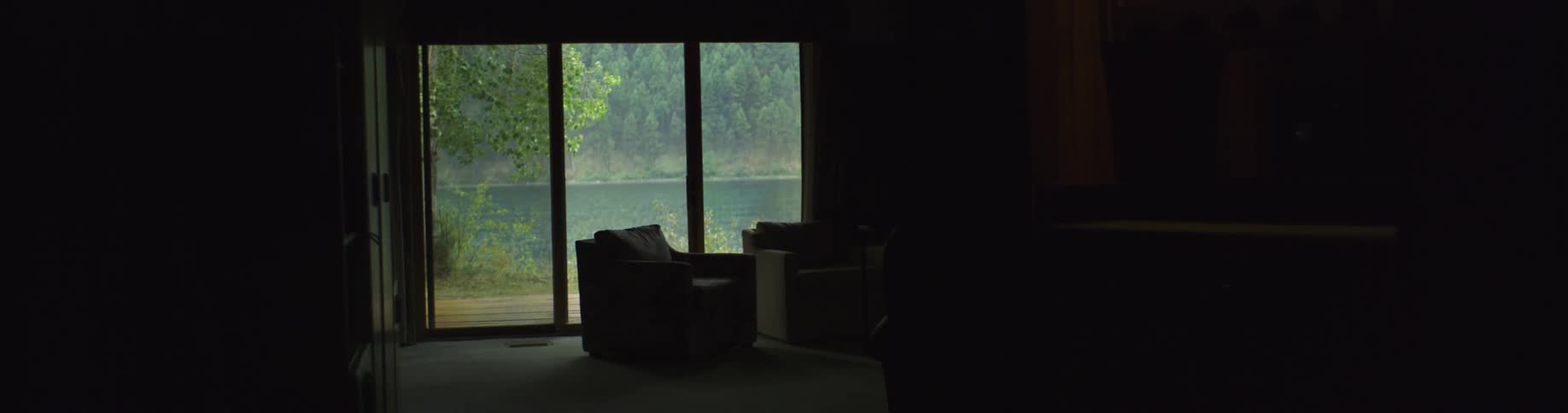 A dark living room illuminated by sunlight from a glass door revealing an outdoor lake