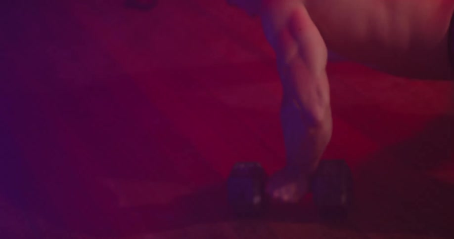 A man doing pushups while alternating a dumbbell between his right and left hand in a room illuminated by a red light