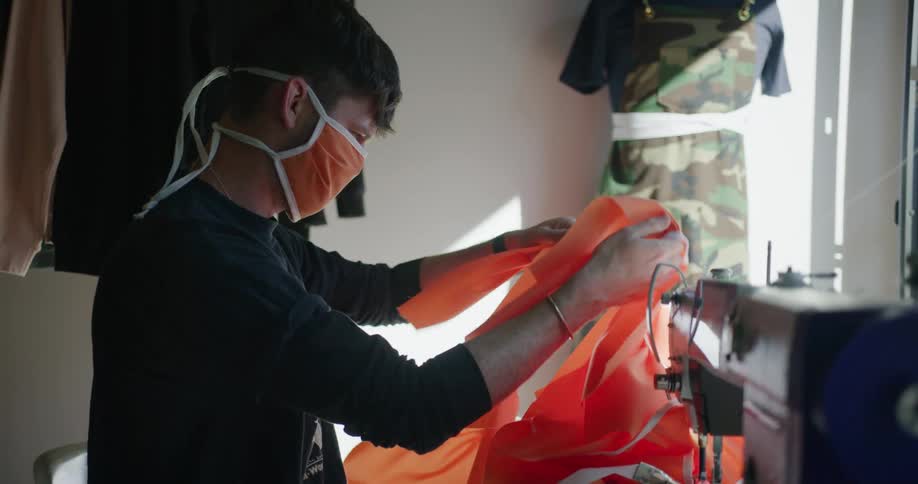 A man wearing a cloth face mask sits at a sewing machine looking through the orange fabric masks he has been sewing