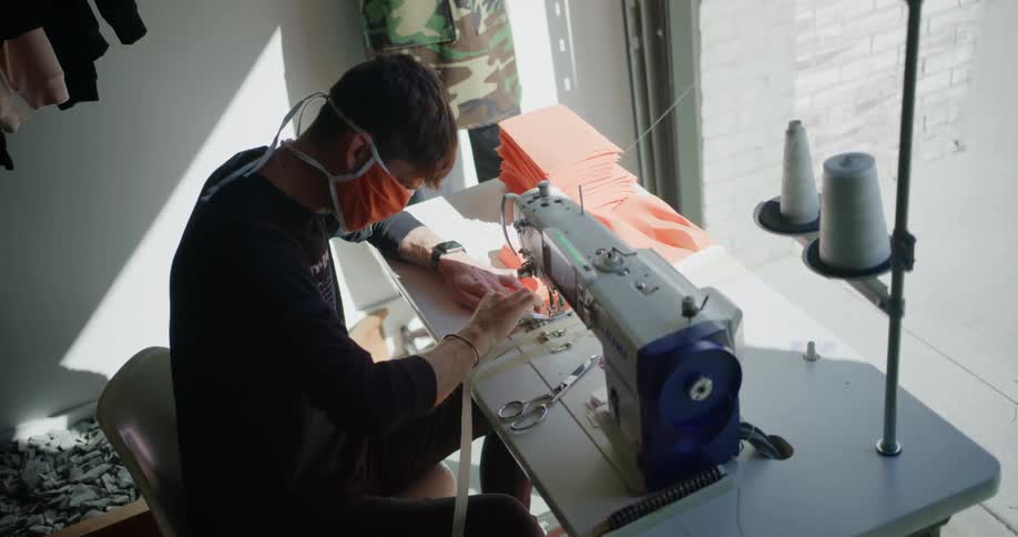 A man sits at a sewing machine in front of a large window making orange cloth masks
