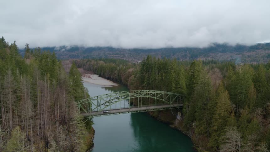 A black car is going over a bridge that is over a river with trees on each side of its banks in winter time