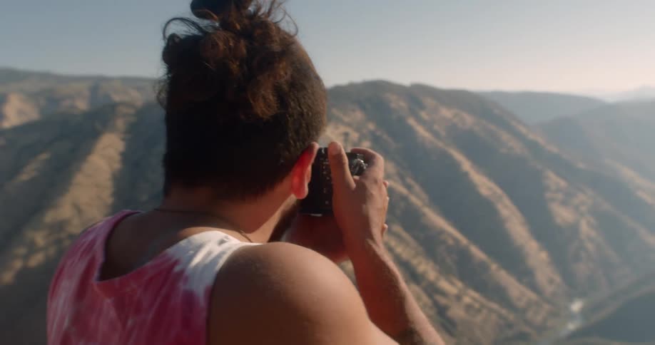 A man in a tie dye tank top takes a photograph of hills canyon and ravine below