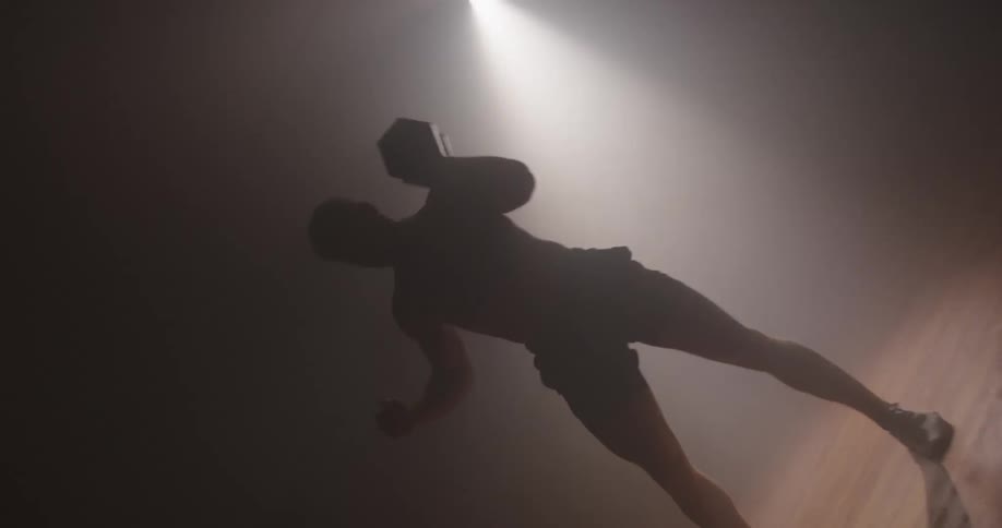 Man lifts weight in shadowy room back lit by spotlight