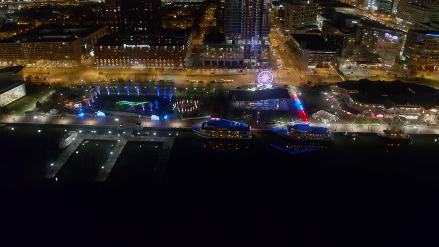 Lights from a ferris wheel and docked boats along the harbor flash and glow along with city lights