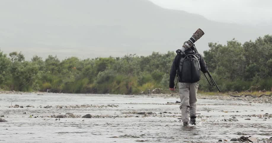 A man carrying a camera and tripod walks through shallow water