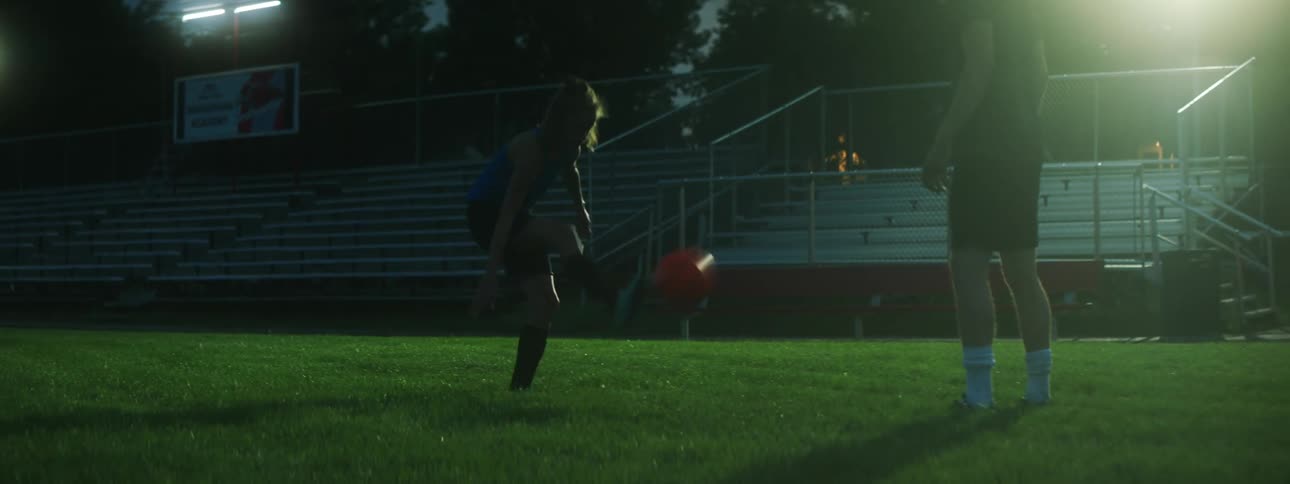 Man and girl practice soccer on field near empty stands at night
