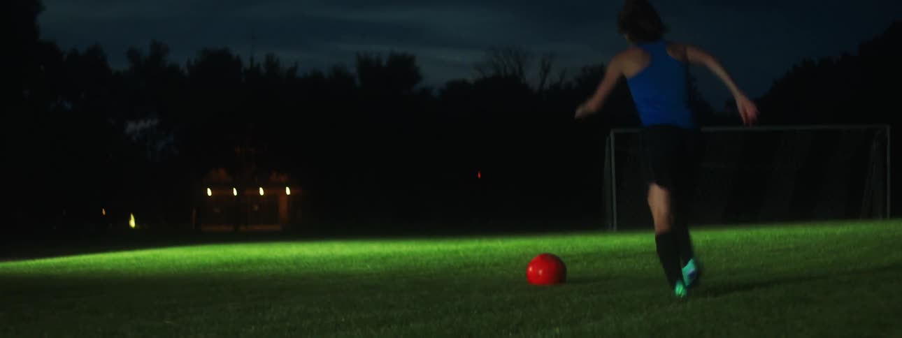 A man and a woman running on a soccer field kicking a ball at each other