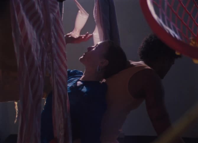 A female dancer leaning backward and a male dancer leaning forward as they stand back to back in a room with hanging shreds of cloth
