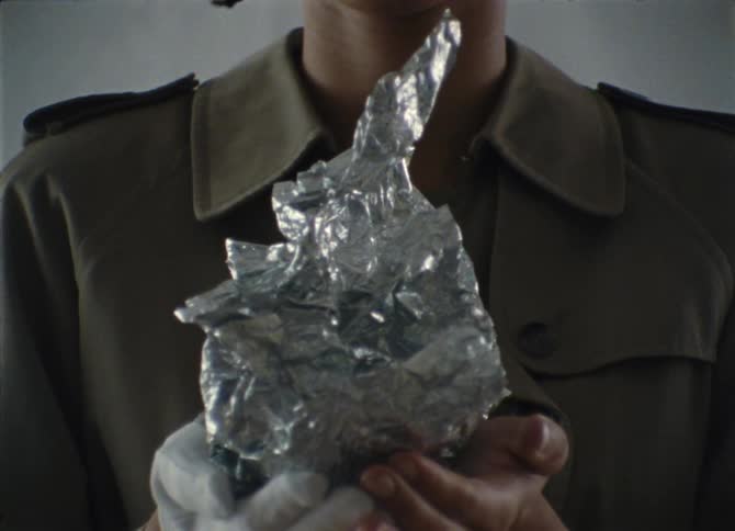 A black woman in a trench coat holds a wad of tin foil in her hands