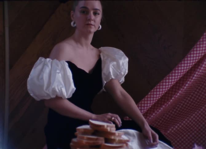 A white woman in a dress sits next to a checkered blanket and a table with bread and candles on it