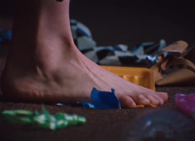 A person walking across a carpet covered in ribbon and wrapping paper steps causing a ribbon to become stuck to the bottom of their foot