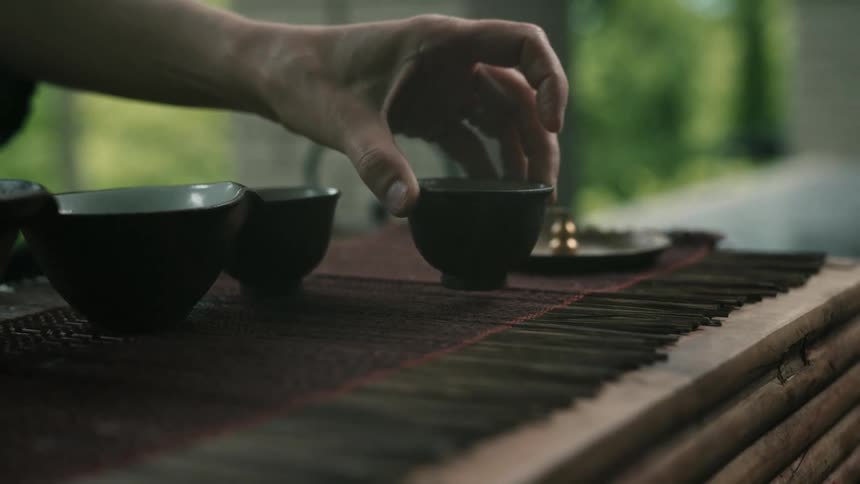 A person sitting at wooden table on the outside of a building moving one of three tea cups to the opposite side of the table