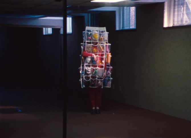 A woman stands in a dark office dressed in a plastic cover lined with pockets that are filled with random toys