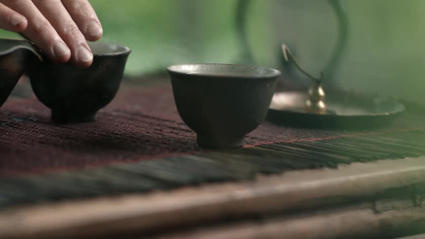 A person sitting at a table placing a steaming cup of tea on the opposite side of the table before picking up a different cup