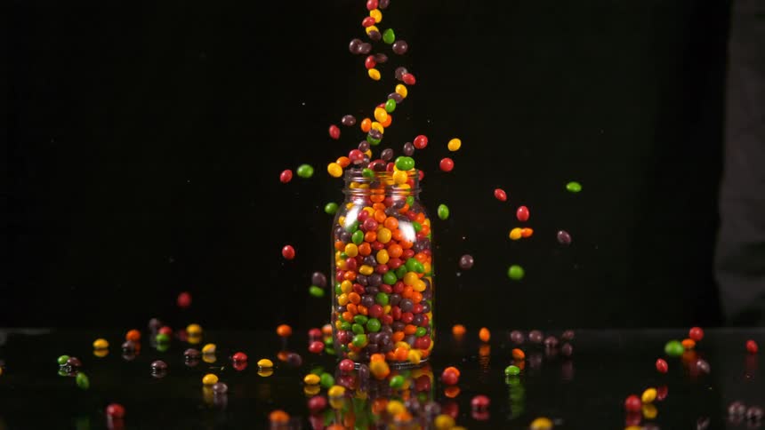 Colorful hard shelled candies are dropped into overflowing glass jar on black glossy surface