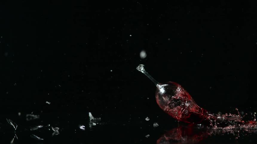 A glass of red wine falls and shatters on a black surface