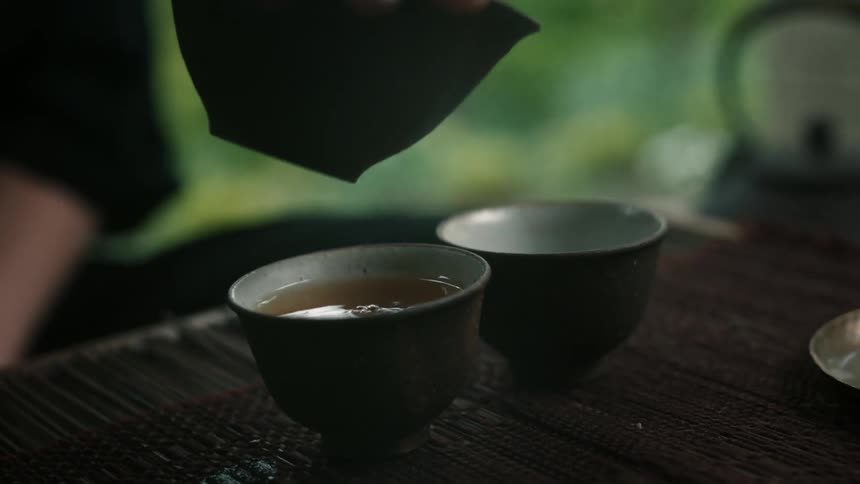 Tea is poured from a teapot into two small tea cups on a bamboo mat