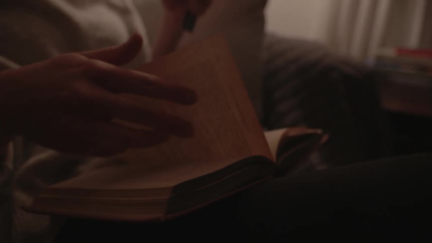 A person reads a book while sitting on a couch in a dimly lit room at home