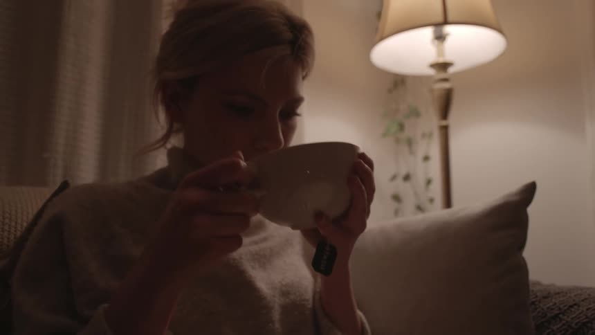 A woman sits on a couch and sips something from a bowl