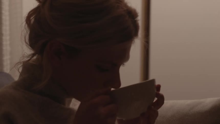 A white woman sits on a couch and drinks from a cup