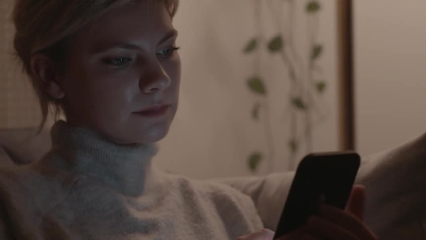 Female scrolling and looking at her cell phone as she pets the cat who jumps up on the sofa she is sitting on and then goes back to scrolling on her phone