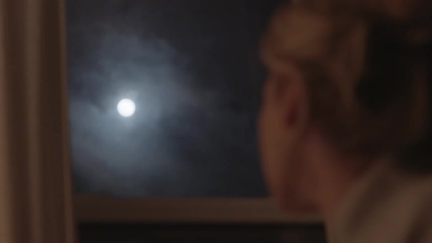 Woman looks out the window at clouds passing over a full moon
