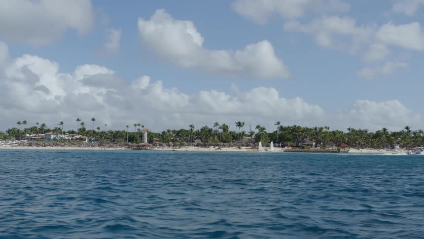 A boat moves along a beach with buildings and palm trees