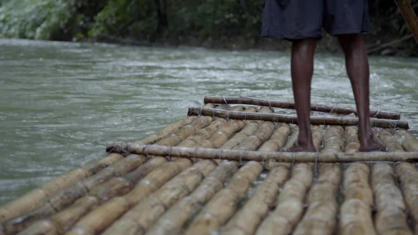 A person standing on a wooden raft guides it down the river with a long oar