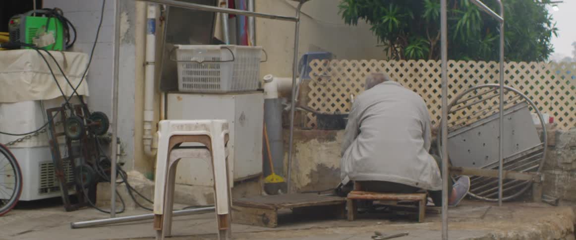 Light flashes and sparks fall as a man uses a torch and tools while sitting on a stool outside