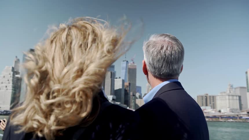 A man and woman stand looking at the ocean and the city skyline on a bright sunny windy day