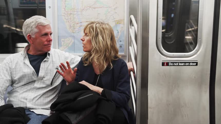 Male and female couple sitting close together on the moving subway train as the female speaks using her hands and the male listens intently