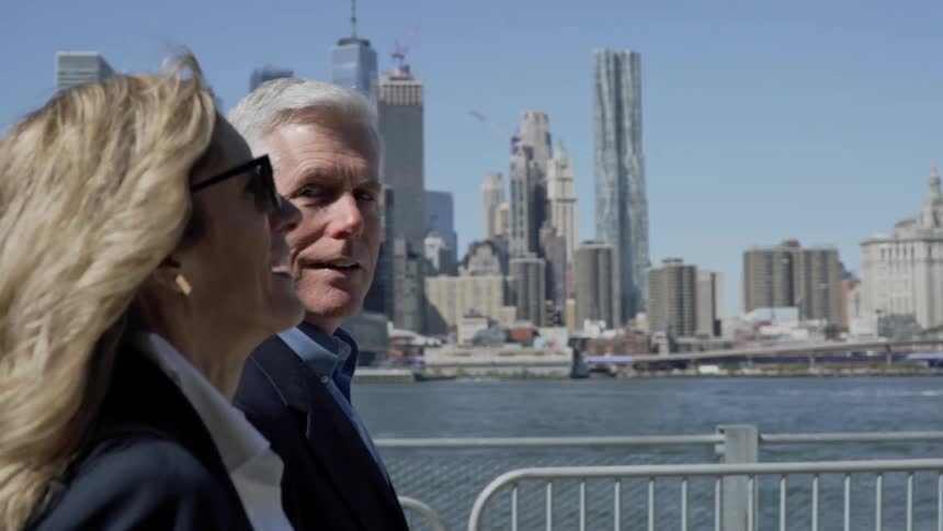 A man and a woman in business attire walk along an urban riverfront