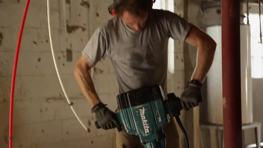 A man strains as he uses a jackhammer to rip up flooring around pipes in a home