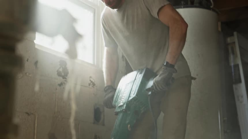 Man in a grey shirt breaks down concrete floor with a jackhammer