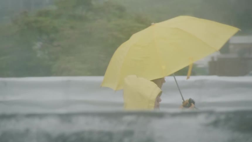 Woman in raincoat walks with woman under yellow umbrella looking at digital camera